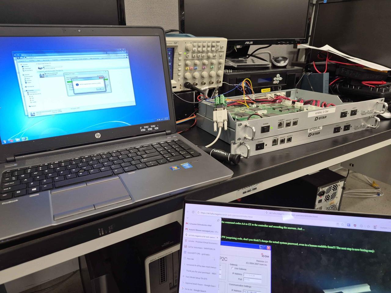 A lab bench with a Windows 7 HP ProBook on the left burning a CD image, an oscilloscope and second monitor center, and two stacked Icom ID-RP2C controllers on the right with the top one open and a custom serial cable running into it. A second monitor at the bottom shows the OK1UHU article on the password procedure.