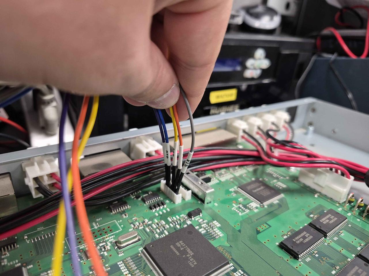 Hand holding a wired-up 6-pin connector. Each lead has a heatshrink label: RTD, RSD, TxD, RTS, etc. The PCB beneath shows large Hitachi-branded ICs.